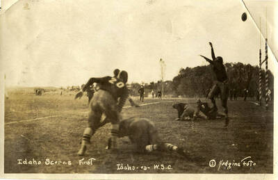 An action shot taken during a football game between the University of Idaho and Washington State College captioned with, 'Idaho Scores First.' The photo captured a man jumping for the ball while other players are on the ground recovering from tackles.