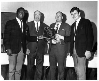 Four man stand together as they hold a clock engraved with Joe Vandal.