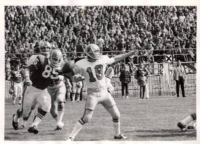 #18 for the Vandals prepares to throw the football as player #85 from the opposing team tries to tackle him from his blind side while the packed audience watches from the bleachers.