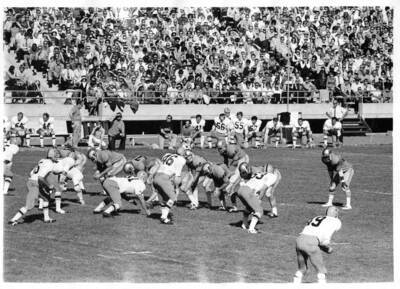 Football players kneel on the sidelines to watch their teammates as they prepare to snap the ball.