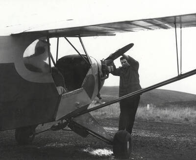 A member of the Vandal Flying Club starts the propeller on a Piper aircraft while another member sits in the cockpit.