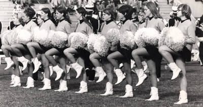 The Pom-Pom Girls perform a cheer on MacLean Field during a football game. The Vandal Marching Band performs in the background behind them.