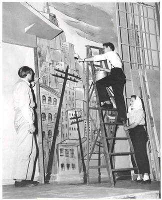 Don Swinney and Kay Thometz watch "Bud" Chamberlain paint a background setting in preparation for the play, 'Sing, Senator, Sing.' An ASUI production written by Geoffrey Coope, Theodore Sherman, & music Hall Macklin (all UI teaching faculty), directed by Jean Collette in the University Auditorium.