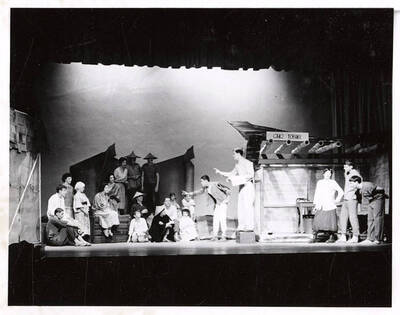Students perform a group scene during a drama production of 'Teahouse of the August Moon.' The actors are costumed as Japanese villagers and American soldiers. Individual performers are identified on the photographs mount.  Directed by Jean Collette in the University Auditorium.