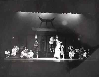 Students perform a group scene during a drama production of 'Teahouse of the August Moon.' Actors are costumed as Japanese villagers and American soldiers. Individual performers are identified on the photograph's mount. Directed by Jean Collette in the University Auditorium.