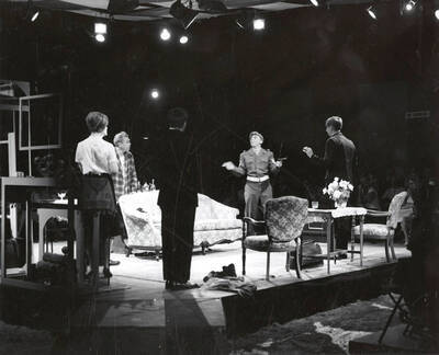 Summer Theatre in the Field House directed by Edmund Chavez. A group of people standing in a circle with one man in uniform holding a gun.