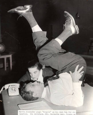 Marvin Alexander plays 'bicycle' on his desk top for relaxation as his secretary, Mrs. Georgette Amos, reminds him of an appointment. Summer Theatre in the Field House directed by Jean Collette.