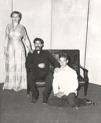 Nina Quick, Marvin Alexander and Albert Alexander. A boy is sitting on the floor. A man is sitting on the chair with his hand on the boy's shoulder. The woman standing next to the chair with her hand on the man's shoulder. Summer Theatre in the Field House directed by Jean Collette.