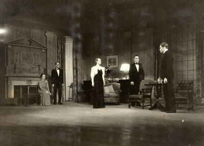 During the drama production of 'Dangerous Corner,' men and women stand on the stage. Cast: Robert Herrick as Gordon Whitehouse, Dorothy Pierce as Freda Chatfield, Eldred Stephenson as Robert Chatfield, Alline King as Olwen Peel, Paul Selby as Charles Stanton, Nina Varian as Betty Whitehouse, Ethlyn O'Neal as Maud Mockridge. Directed by Fred Blanchard.