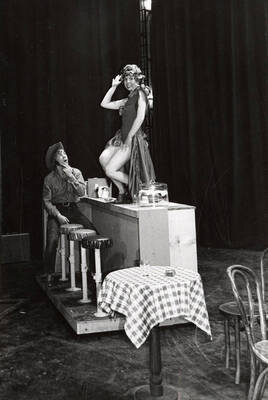 A woman stands on a counter, while a seated man looks on during a drama production. Directed by Forrest Sears for Summer Repertory Theatre in the Performing Arts Center (Hartung Theater).