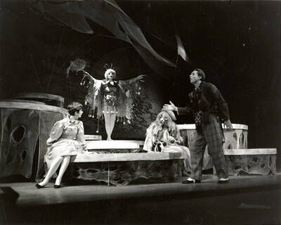 Scene from the University of Idaho drama production of "The Man in the Moon." A woman can be seen dressed in costume with her arms spread. Three other actors can be seen conversing with the woman. Directed by John Naples in the University Auditorium.