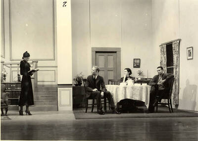 Group scene from the University of Idaho drama production of "Three Cornered Moon." Three actors can be seen sitting around a table, conversing with a woman. Cast members include Helen Wilson as Elizabeth Rimplegar, Erma Lewis as Mrs. Eimplegar, Aldrich Bowler as poet, Jean Ricker as Kitty, Burnell Baker as Dr. Stevens, and Catherine Bjornstad as Jenny. Directed by Fred Blanchard, Technical Director Theodore Prichard in the University Auditorium.