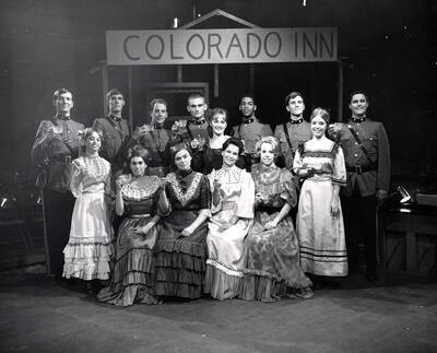 Group photo of the cast from the University of Idaho drama production of "Little Mary Sunshine." Directed by Edmund Chavez for Summer Theatre in the Field House. Women are sitting and standing in front, wearing dresses, and men stand in the back, also in costume. The cast hold raised glasses under a sign that says 'Colorado Inn.'