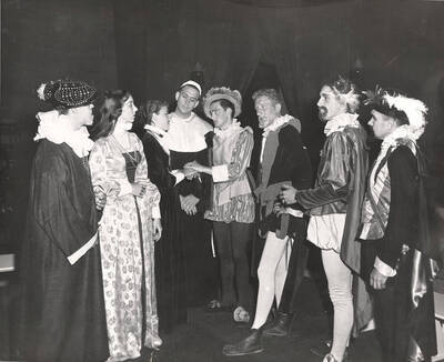 Group scene during the University of Idaho drama production of "Much Ado About Nothing." Directed and designed by Edmund Chavez for Summer Theatre in the Field House. This production was directed by Edmund Chavez. Members of the cast are dressed in costume and all standing around talking with each other.
