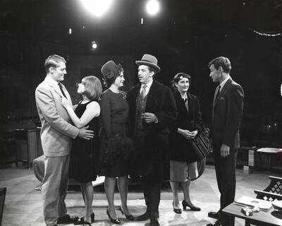 Scene during the University of Idaho drama production of "Come Blow Your Horn." Directed by Edmund Chavez for Summer Theatre in the Field House. A group of people can be seen standing on stage in costume and conversing with each other.