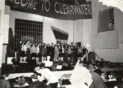 Scene from the ASUI production of "Gee-Eyes Right." The cast members can be seen standing on stage under a sign that reads 'Welcome to Clearwater'. Directed by Jean Collette, written Ted Sherman and Geoffrey Cope, music by Hall Macklin, costume advisor Marion Featherstone.