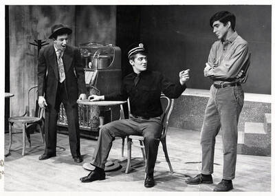 Scene from the ASUI Dramatic production of "Andorra." This production was directed by Jean Collette. One man can be seen sitting at a table, talking with two other men.
