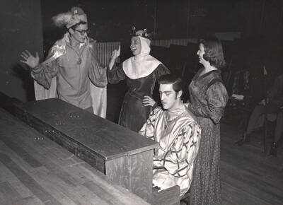 Clyde Winters as Osric, Margaret Magee as Gertrude, Charles LaFollette as Rosencrantz and Jean Bales as Lady in Waiting stand around a piano during a break from rehearsal of 'Hamlet.'  Directed by Jean Collette, costumes by Joan Coble (Judy Chavez) in the University Auditorium.