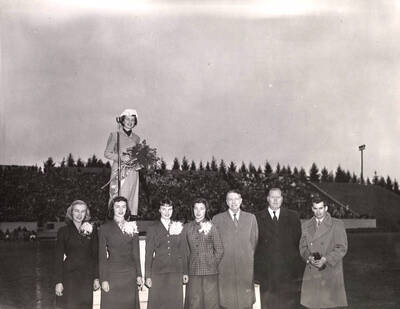 Homecoming queen Leah Jensen holds a bouquet on a dais. Pictured in front of the dais from left to right: Jackie Taylor, Betty Westerberg, Barbara Thurston, Shirlie Vorous, Governor Len Jordan, Larry Pugh (Alumni President), and Hyde Jacobs, ASUI student body president.