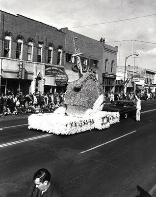 An unidentified float drives through the Homecoming parade in front of Wards, Farmers Insurance, Creighton's, Bjorklund Hardware and Norge Hanson Electric.