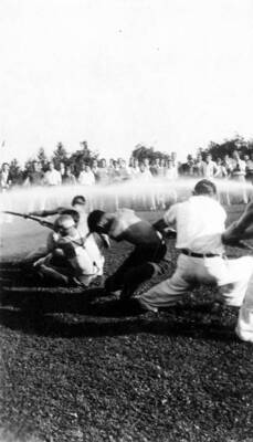A crowd watches as a group of men playing tug-of-war duck under a stream of water.