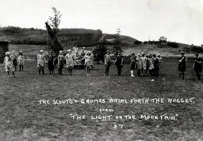 People pose holding hands, mostly in gnome and scout garb, for the Light on the Mountains pageant. Caption reads: 'The Scouts & Gnomes Bring Forth the Nugget. From 'The Light on the Mountain.' #7.'