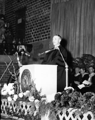 Governor Len Jordan stands at a podium giving a speech in Memorial Gymnasium for University of Idaho's Commencement.