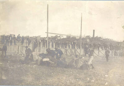 Several players pile up in the end zone. Fans can be seen standing on the wooden fence in the background.