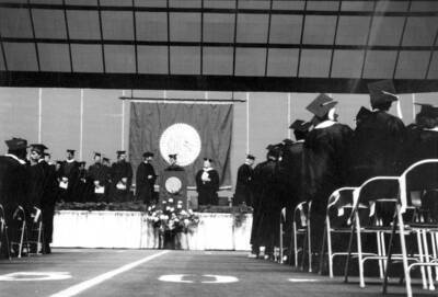 A ground level photograph of the stage and podium shows faculty and regents waiting for the beginning of the Commencement ceremony.