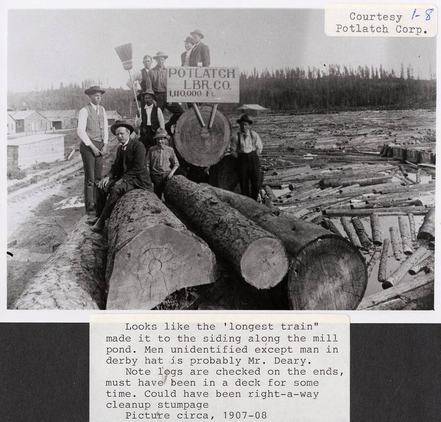 The longest train made it to the siding along the mill pond. A group of men can be seen sitting and standing on a stack of longs. A signs saying 'Potlatch LBR. Co. 1,110,000 Ft.' is also on the stack of logs.