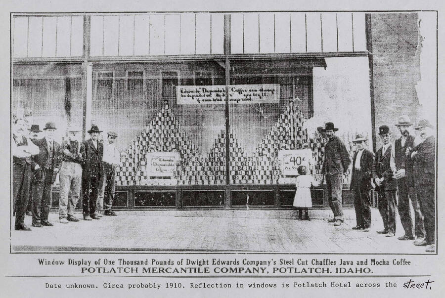 A photograph of a window display of one thousand pounds of Dwight Edwards Company's steel cut chaffles java and mocha coffee at the Potlach Mercantile Company in Potlatch, Idaho. There is a reflection of the Potlatch Hotel across the street. Circa. 1910