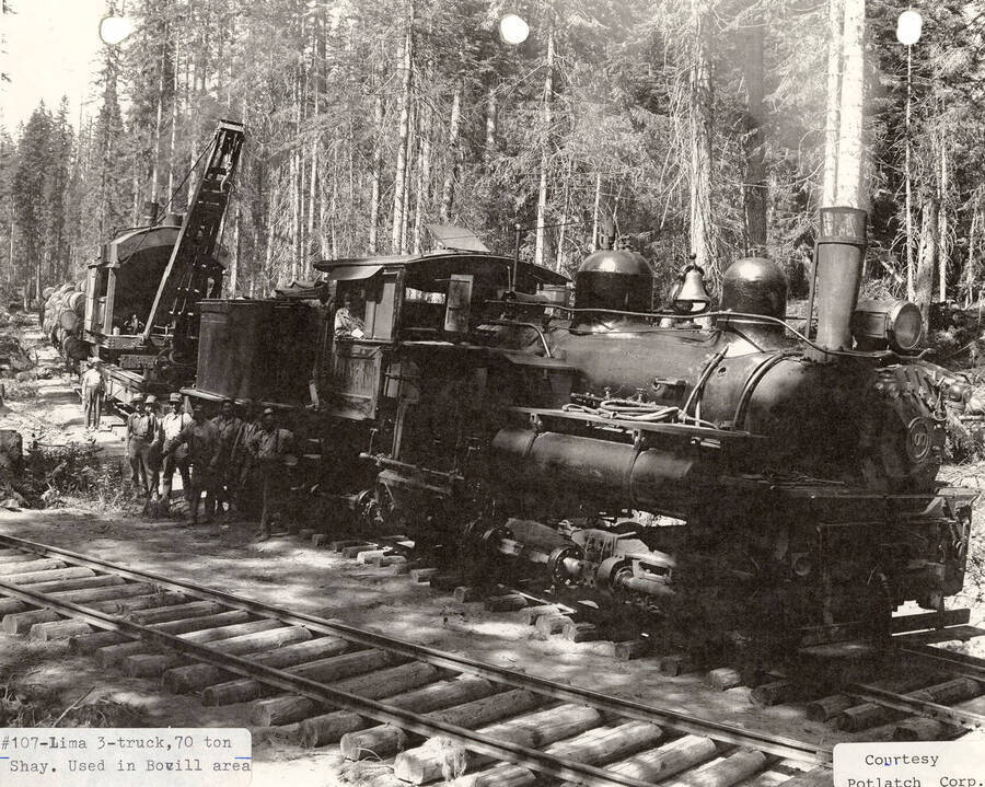 View of a 70 ton Shay locomotive that was used in the Bovill area. A few men are standing next to the engine and a piece of equipment can be seen hauling logs behind the engine.