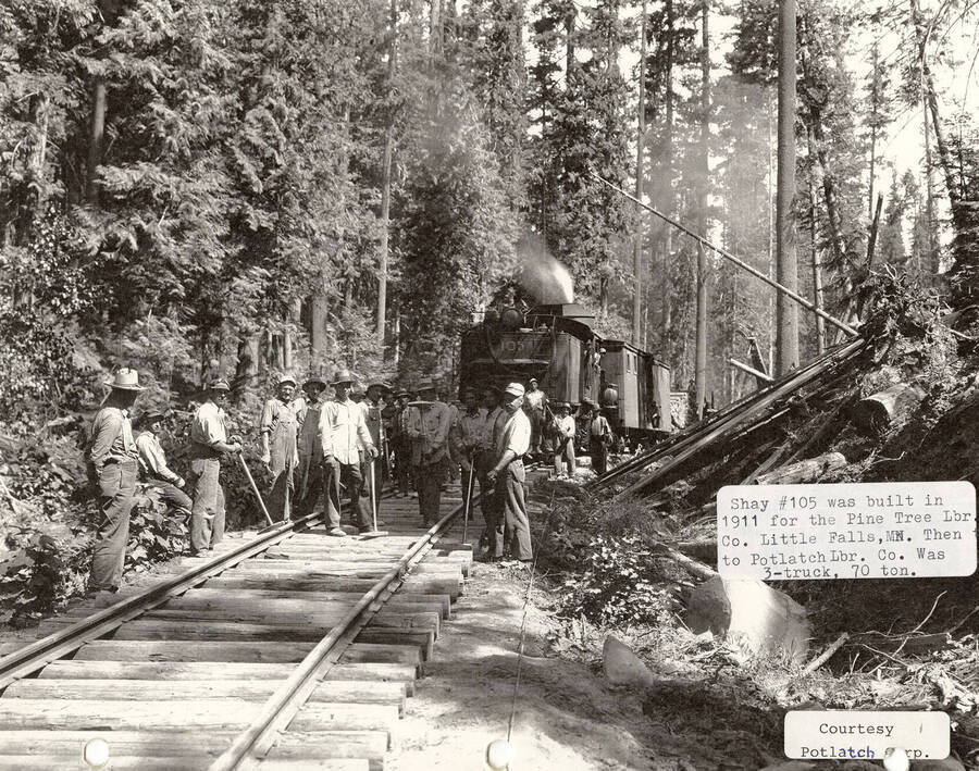 View of the Shay locomotive #105, which was built in 1911 for the Pine Tree Labor Co. in Little Falls, MN. The engine was later given to the Potlatch Lumber Co. Many men can be seen standing in front of the engine on the railroad tracks.