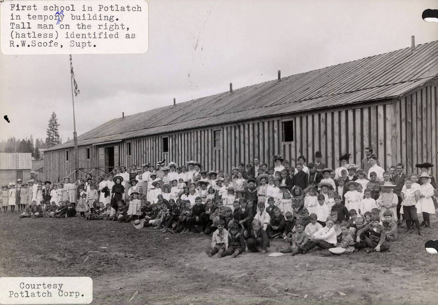 A photograph of the students and temporary building of the first school in Potlatch, Idaho. The Superintendent  R. W. Scofe has been identified as the tall man on the right without a hat.
