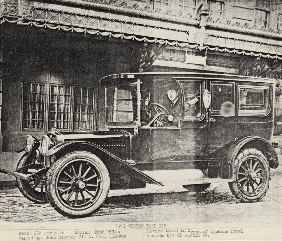 Fred Wilks behind the wheel of the Winton Taxi cab while it is parked in front of the Spokane Hotel.  In 1917 the taxi cab's owner was John Mahoeny and the fare was 25 cents per mile.