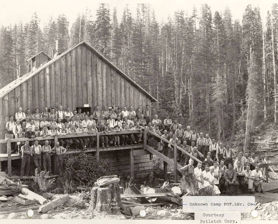 Group photo of the men at a Potlatch camp. The men can be seen standing and sitting on the deck and stairs of a building on the camp.