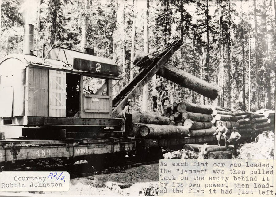 View of a piece of equipment loading flat cars with logs. A few men can be seen standing on and around the stacks of logs.