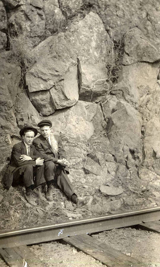 A photograph of two men between a wall of rocks and railroad tracks.