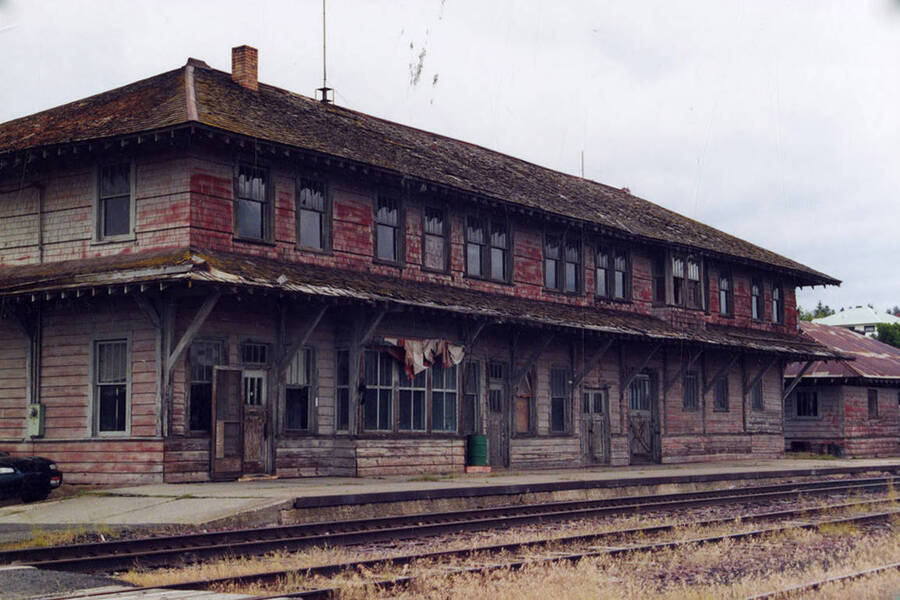 View an old building. A car can be seen parked on the side of the building and a railroad track runs in front of it.