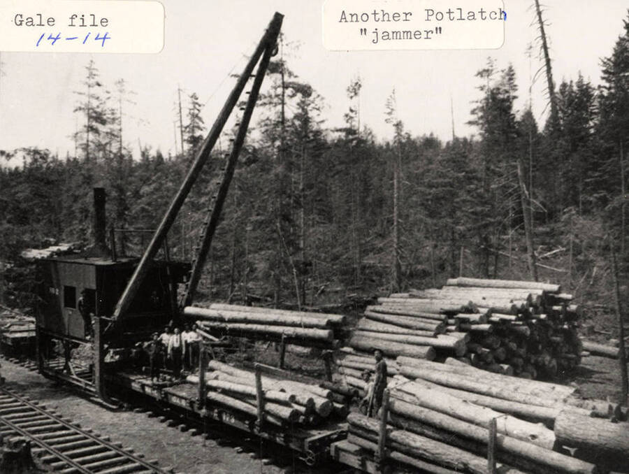 A few men standing on the 'jammer' that is stacking logs on flat cars.