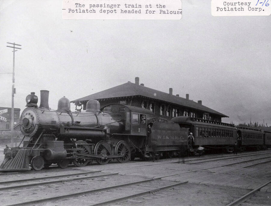 View of a passenger train at the Potlatch depot headed for Palouse. Men can be seen standing on the sides of the train and a few people can be seen in the windows of the train.