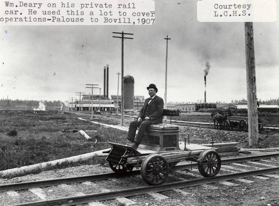 William Deary sitting on his private rail car. He used this often to cover operations between Palouse and Bovill. A few buildings and some horses pulling wagon can be seen in the background.
