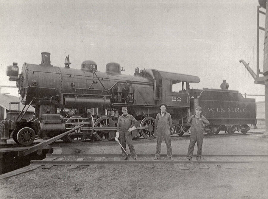 View of a No. 22 locomotive. Three men can be seen standing on the railroad tracks in front of the locomotive.