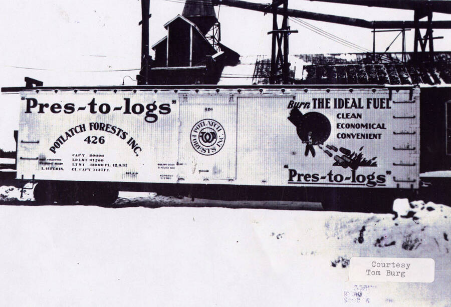 View of a railroad car sitting at a railroad station. The side of the car advertises 'Pres-to-logs' through the Potlatch Forests Inc., which talks about burning the ideal fuel.