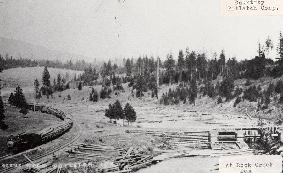 View of the Rock Creek Dam near Potlatch, Idaho. A locomotive hauling stacks of logs can be seen on the railroad tracks to the left of the dam.