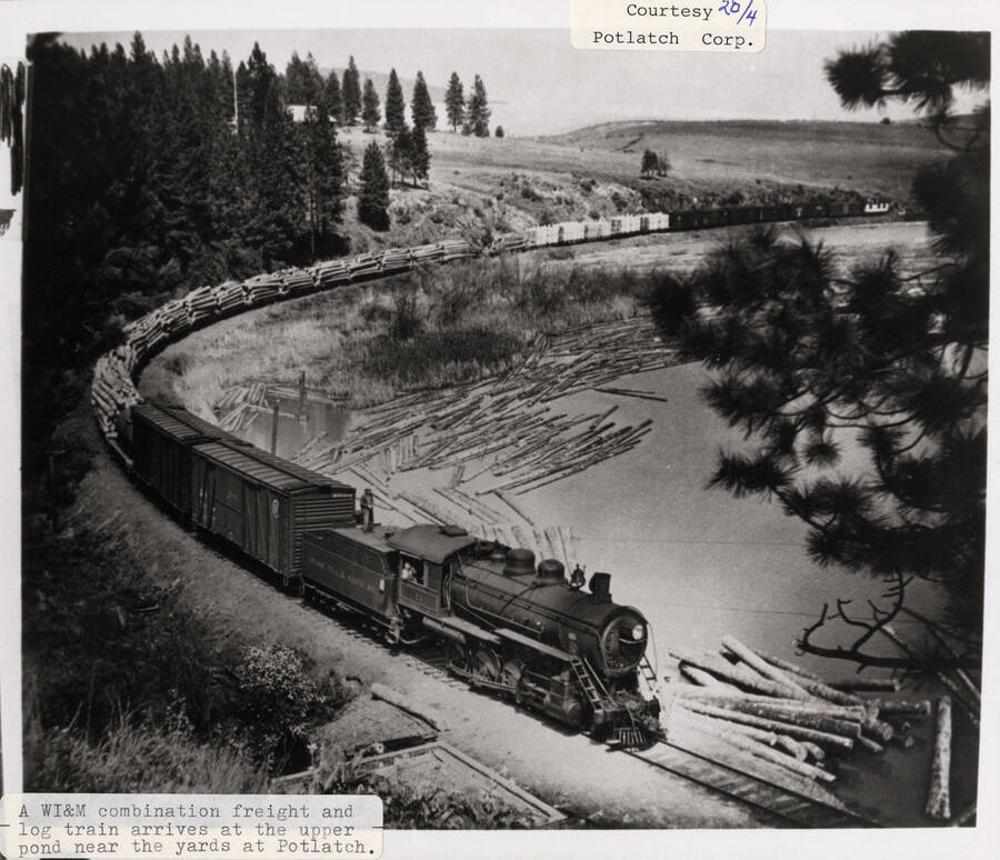 View of a Washington, Idaho, and Montana Railroad combination freight and log train hauling stacks of logs. The train is arriving at the upper pond near the yards at Potlatch, Idaho.