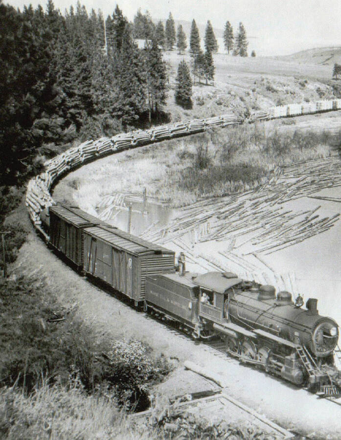 View of a locomotive pulling tacks of logs. A man can be seen sitting next to the window of the locomotive.
