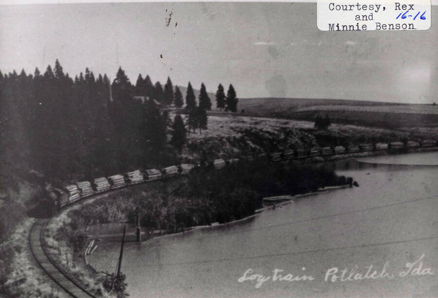 View of a locomotive pulling stacks of logs in Potlatch, Idaho. The railroad can be seen winding around a body of water on one side and a group of trees on the other.