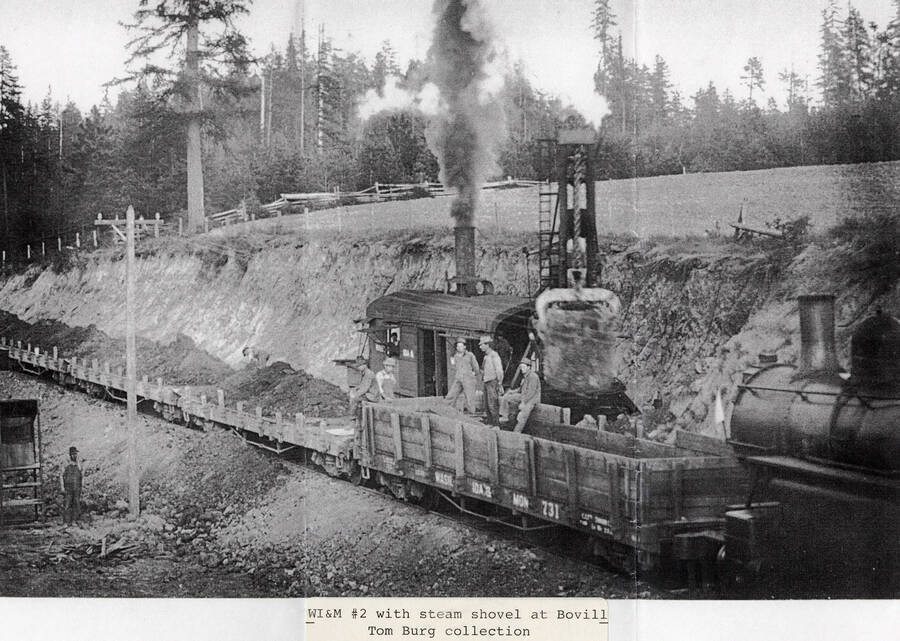 The WI&M No. 2 locomotive wuith a steam shovel. A few men can be seen sitting on one of the railroad cars and other men can be seen standing around next to dirt piles.