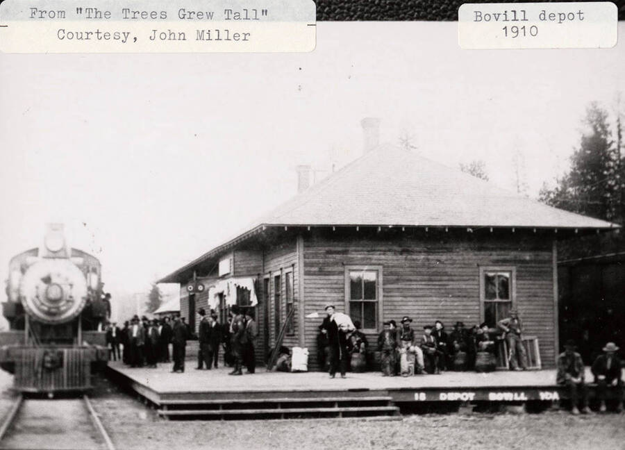View of the Bovill depot. People can be seen sitting on the deck of the depot as a locomotive pulls into the station.